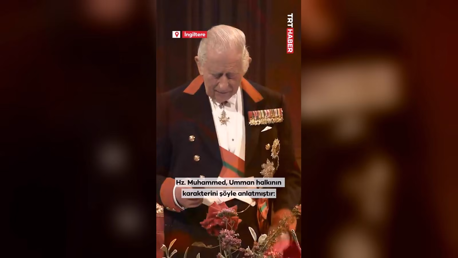 Kırmızı-yeşil kuşaklı, madalyalı üniformalı bir erkek elindeki kağıda bakarak okuyor; önünde çiçek düzenlemesi var.