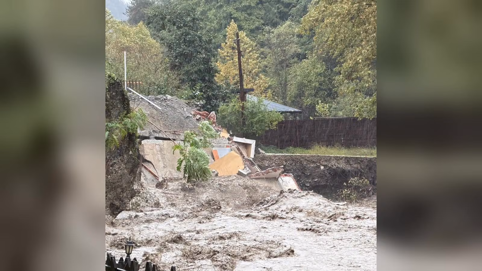 Yağmur altında çamurlu sel suyu hızla akıyor; kıyıdaki duvar ve bir yapı yıkılmış, arkada ağaçlar var.