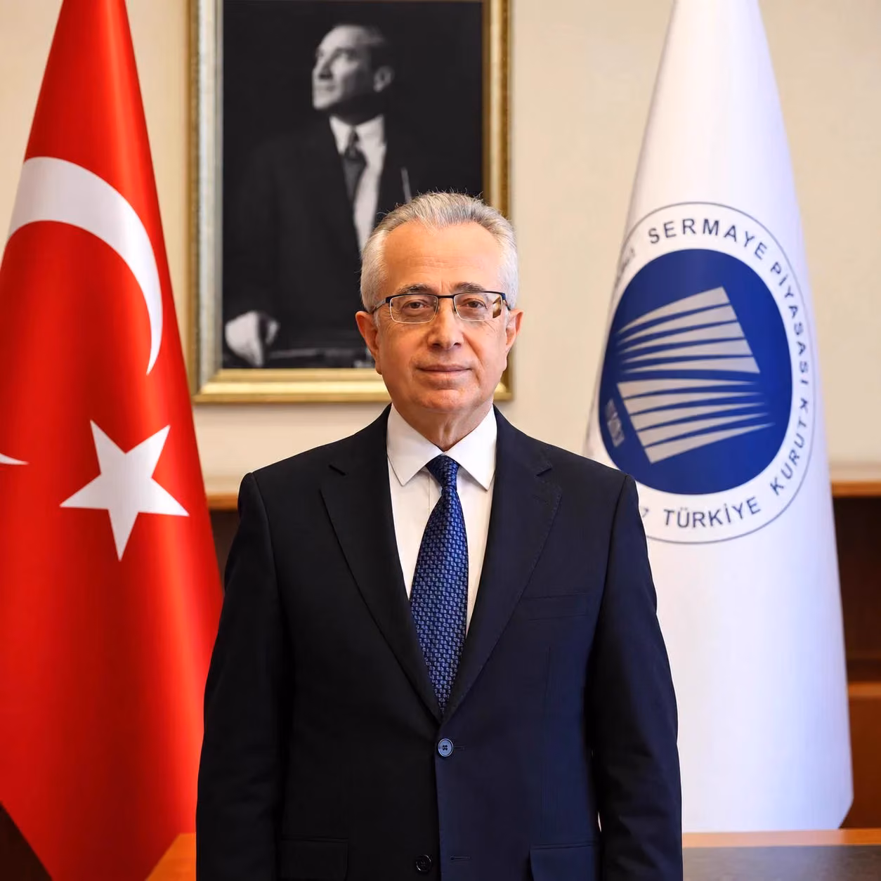Gözlüklü, gri saçlı, lacivert takım elbiseli bir adam; Türk bayrağı ve SPK logolu bayrak önünde, arkada Atatürk portresi var.