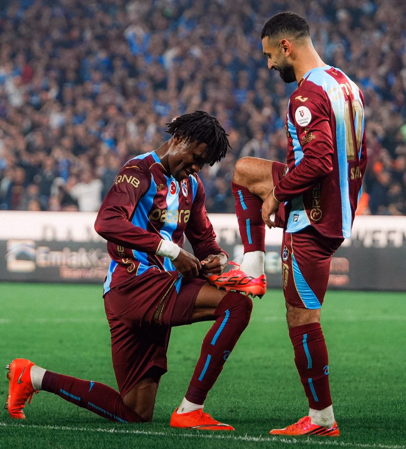 Bordo-mavi formalı iki futbolcu sahada; biri diz çökmüş, kırmızı krampon bağcığını bağlıyor.