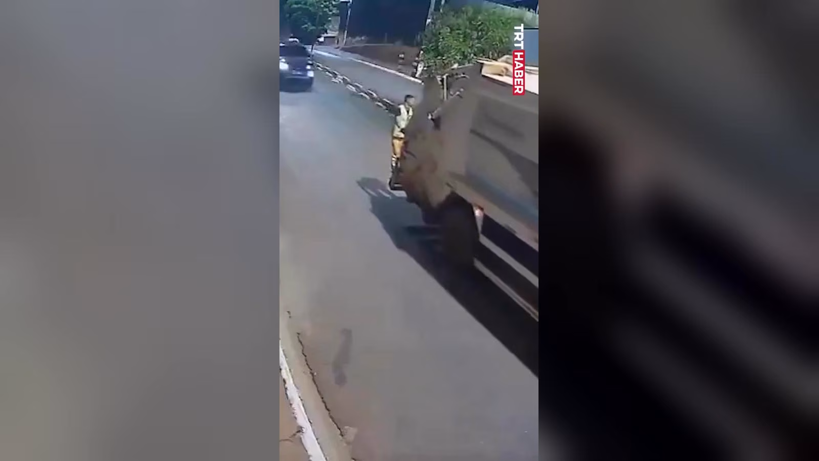 Sarı kıyafetli bir işçi, yolda gri çöp kamyonunun arka tarafının yanında duruyor. Solda bir araç görünüyor.