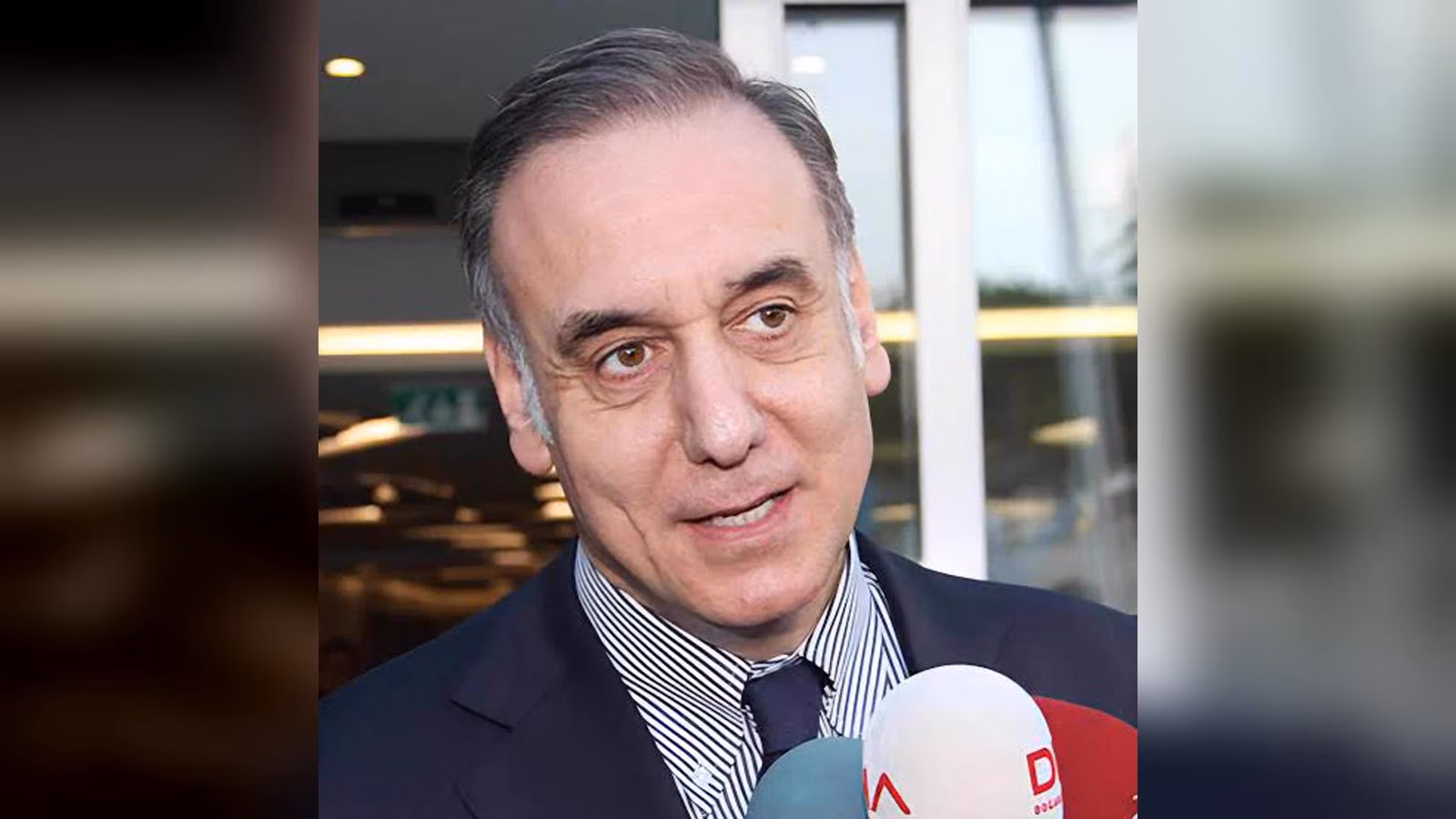 Koyu lacivert takımlı, çizgili gömlek ve koyu kravat takan bir adam, önündeki mikrofonlara konuşuyor.