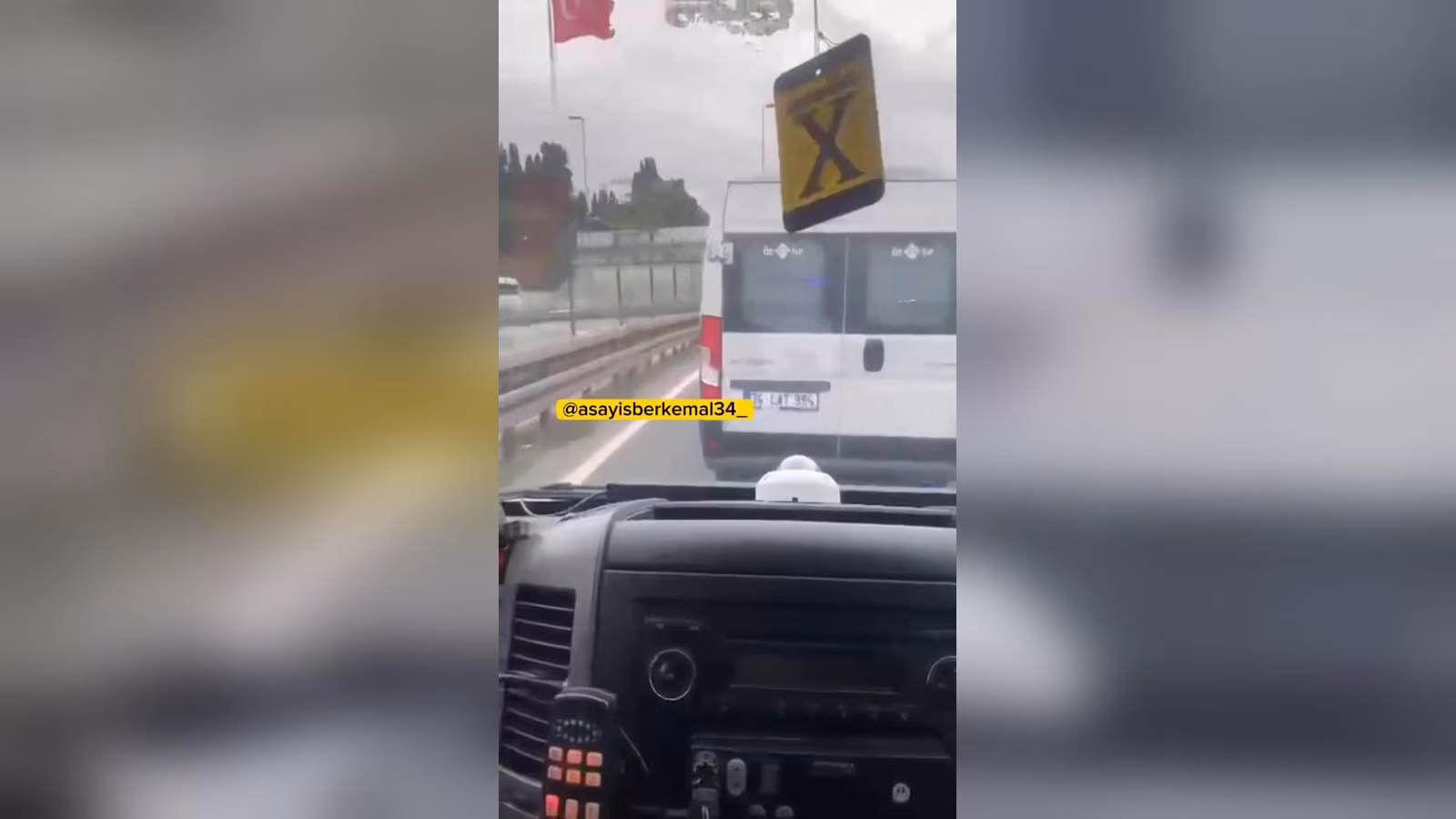 Yağmurlu bir yolda, araç içinden görüntü: önde beyaz bir servis minibüsü var, ön camın yanında sarı X işaretli bir tabela sal