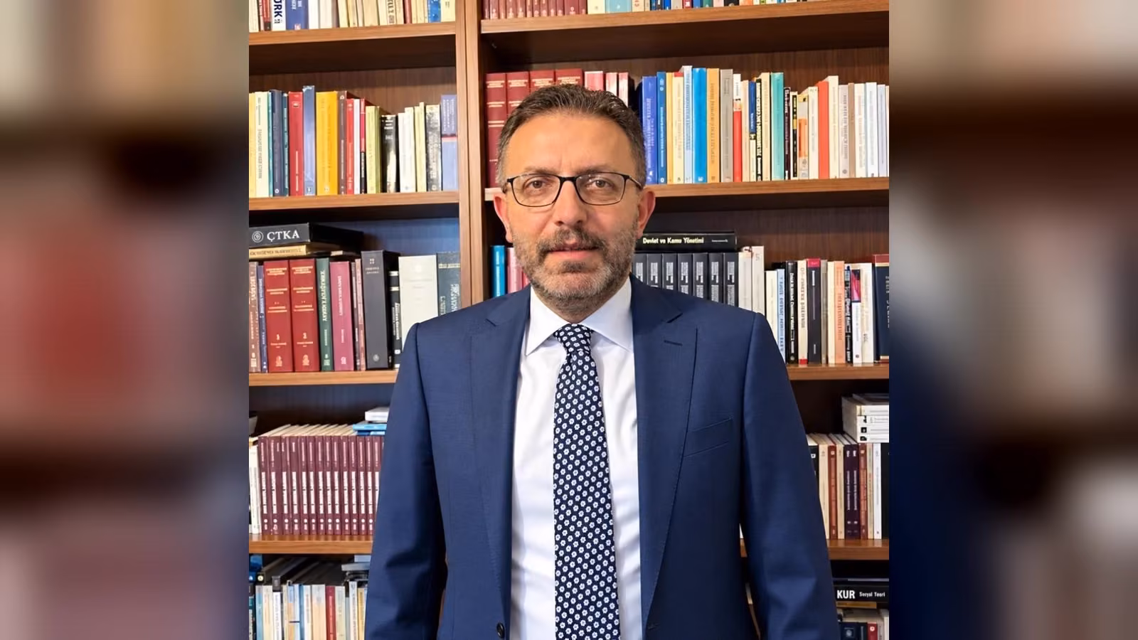 A bespectacled man in a navy suit, white shirt and navy patterned tie, standing in front of a bookshelf.