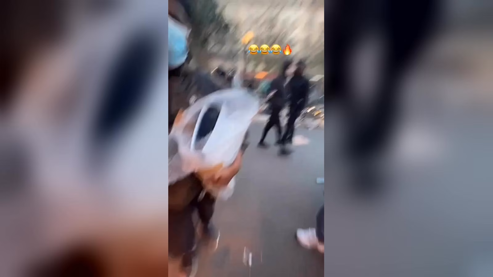 Sokakta beyaz bir oyun konsolu kutusunu tutan bir kişi; arkada koyu kıyafetli kişiler ve bulanık bir görüntü.