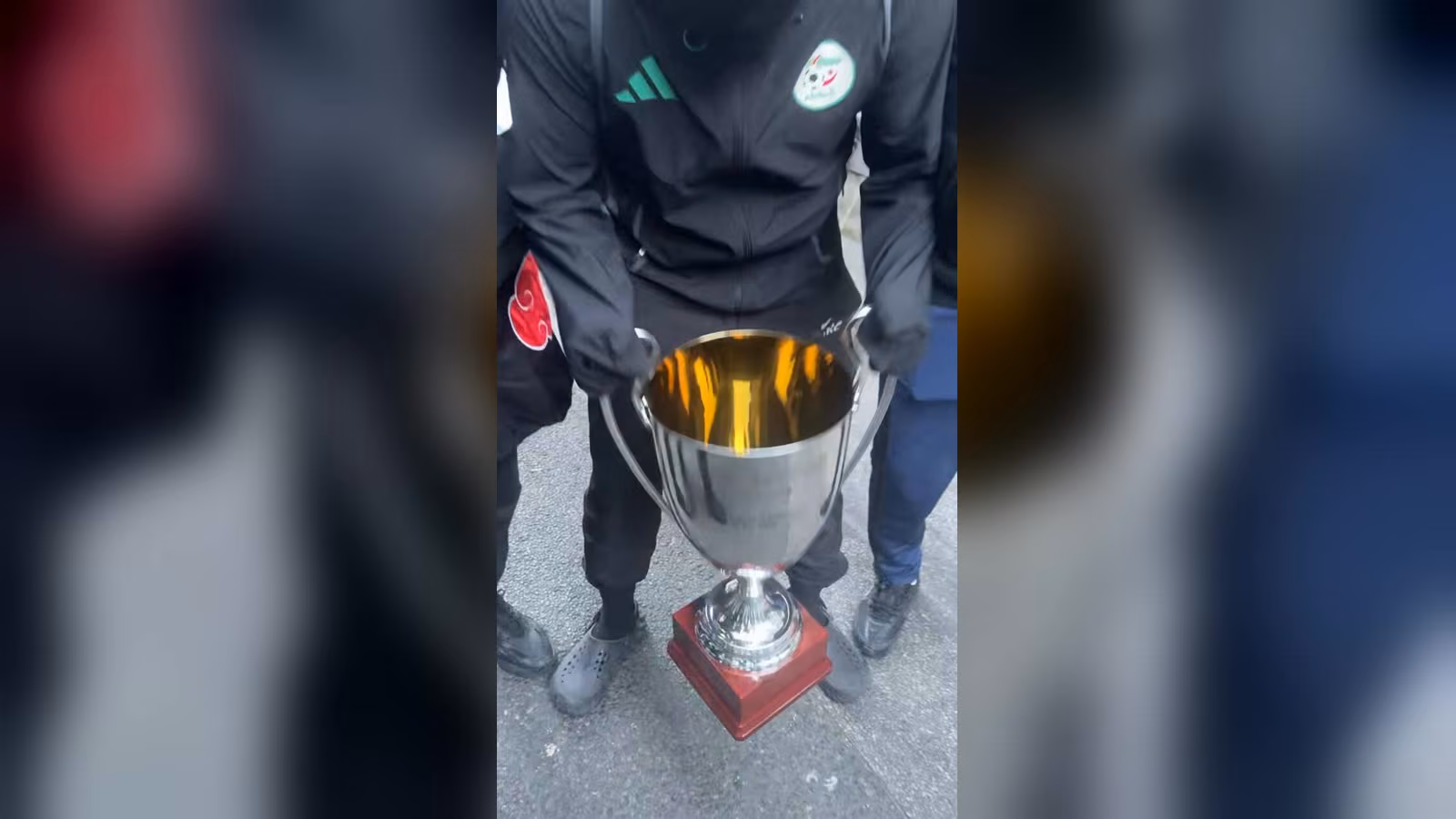 Siyah eşofmanlı bir kişi, kırmızı ahşap tabanlı gümüş renkli bir kupayı asfalt zeminde tutuyor.