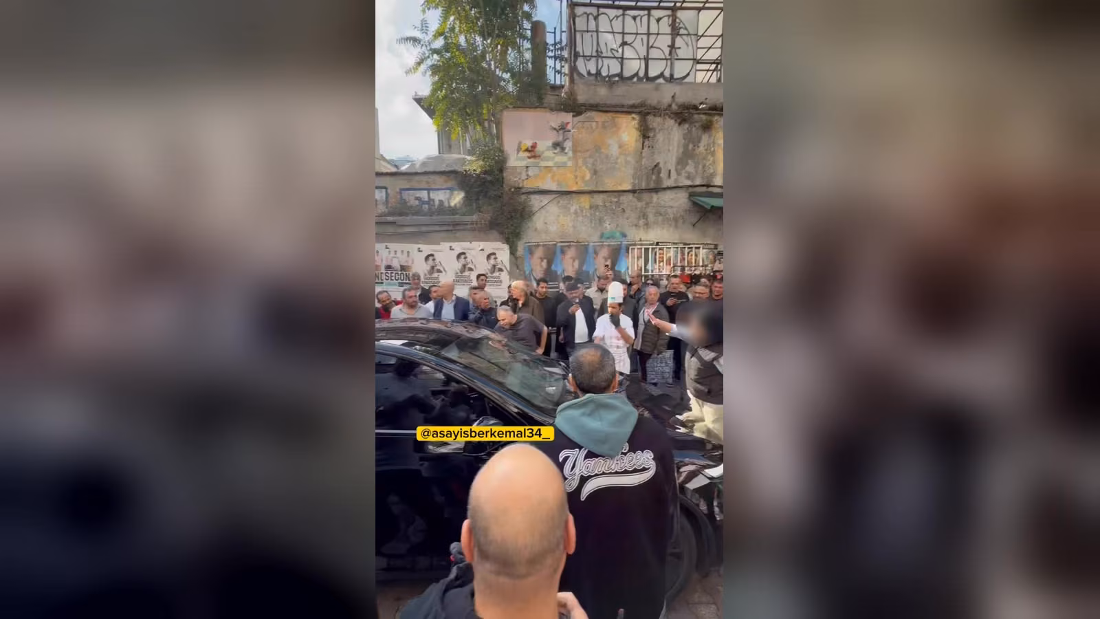 Dar bir sokakta siyah aracın etrafında toplanmış kalabalık; beyaz şapkalı bir adam ve kaputun yanındaki bir kişi.