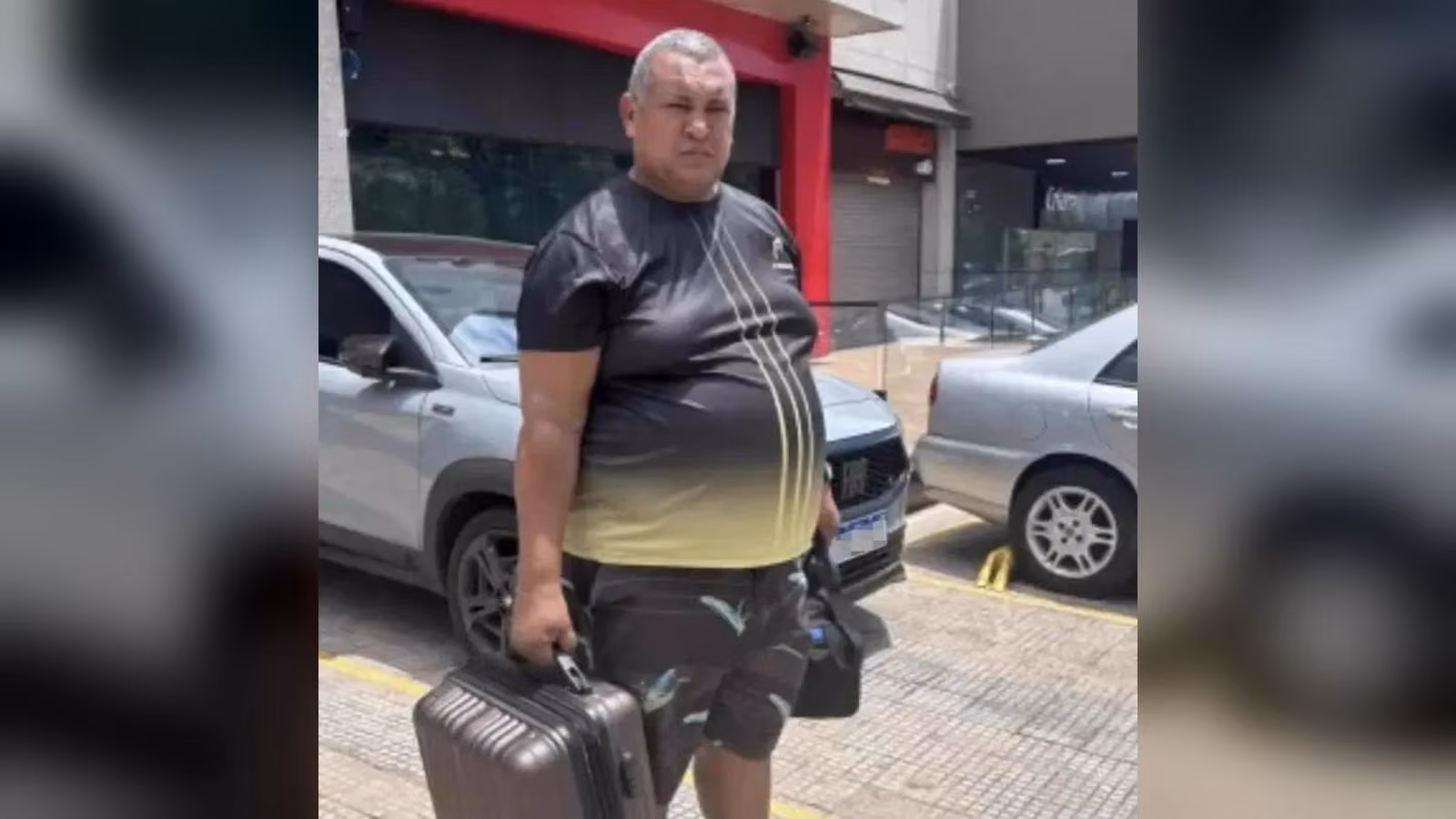 Gri bavulunu ve siyah çantasını taşıyan, koyu renk tişörtlü ve desenli şortlu kilolu bir adam kaldırımda duruyor.
