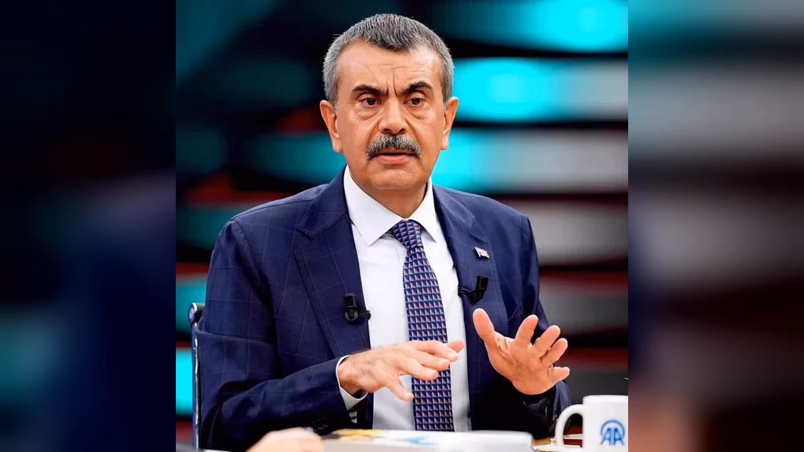 Gri saçlı, bıyıklı, lacivert takım elbiseli ve desenli kravatlı bir erkek, bir televizyon stüdyosunda iki elini açarak konuşu