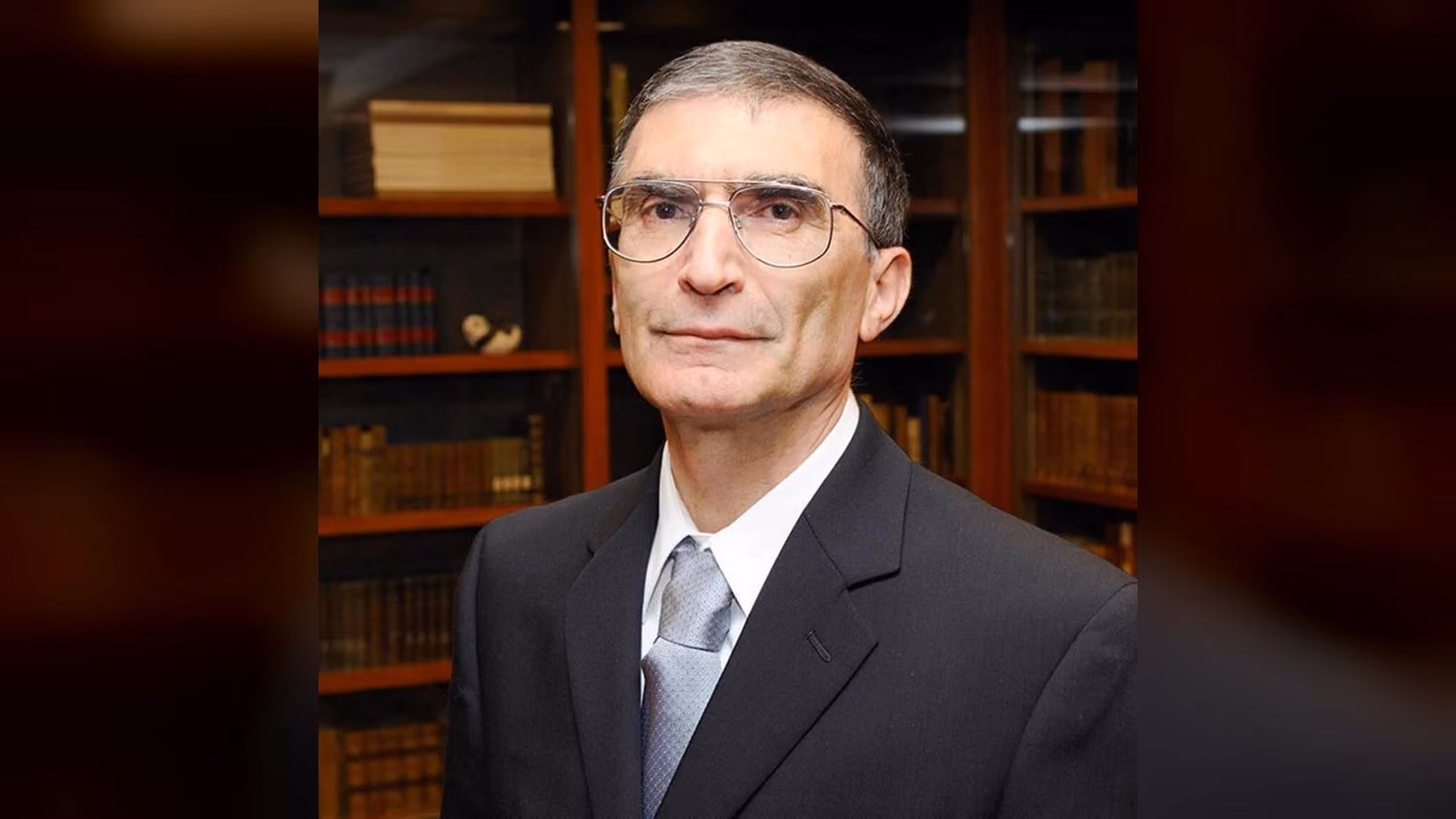 Koyu renkli takım elbiseli, gözlüklü, gri saçlı bir erkek kitaplık önünde kameraya bakıyor.