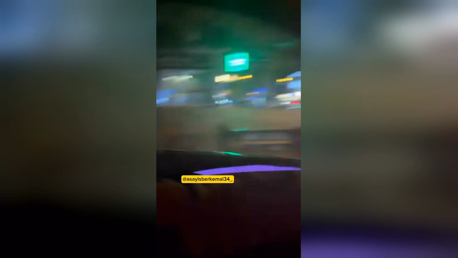 Gece vakti araç içinden çekilmiş, bulanık bir şehir caddesi; yeşil tabela ve ışıklı dükkan vitrinleri görünüyor.