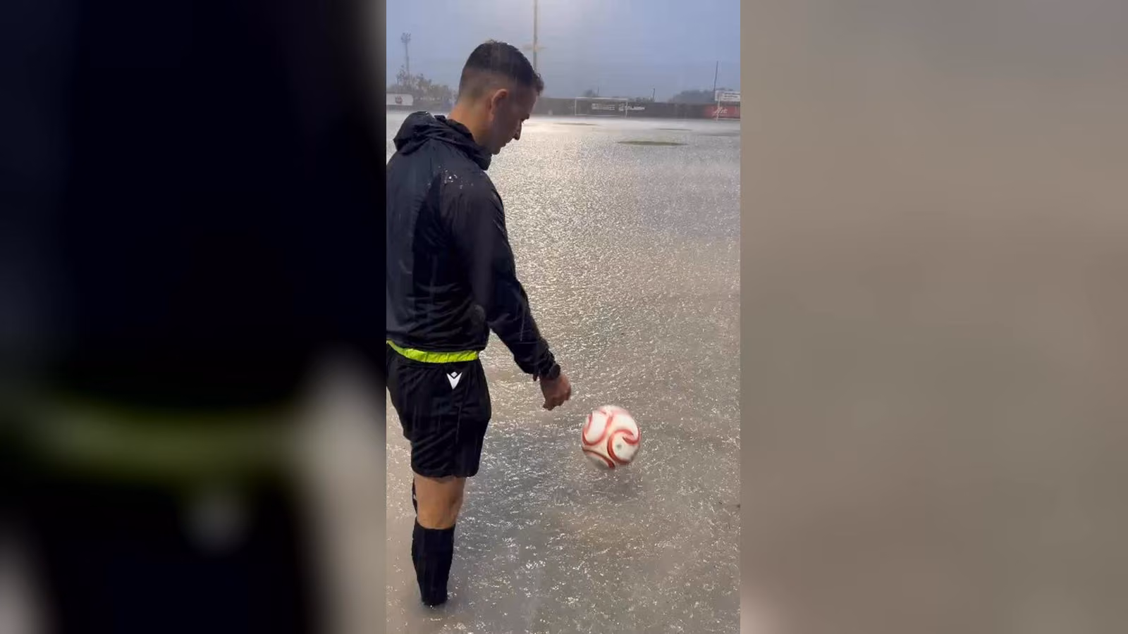 Yağmurla su dolan futbol sahasında, siyah kıyafetli bir hakem suyun içinde topu ayağıyla çeviriyor.