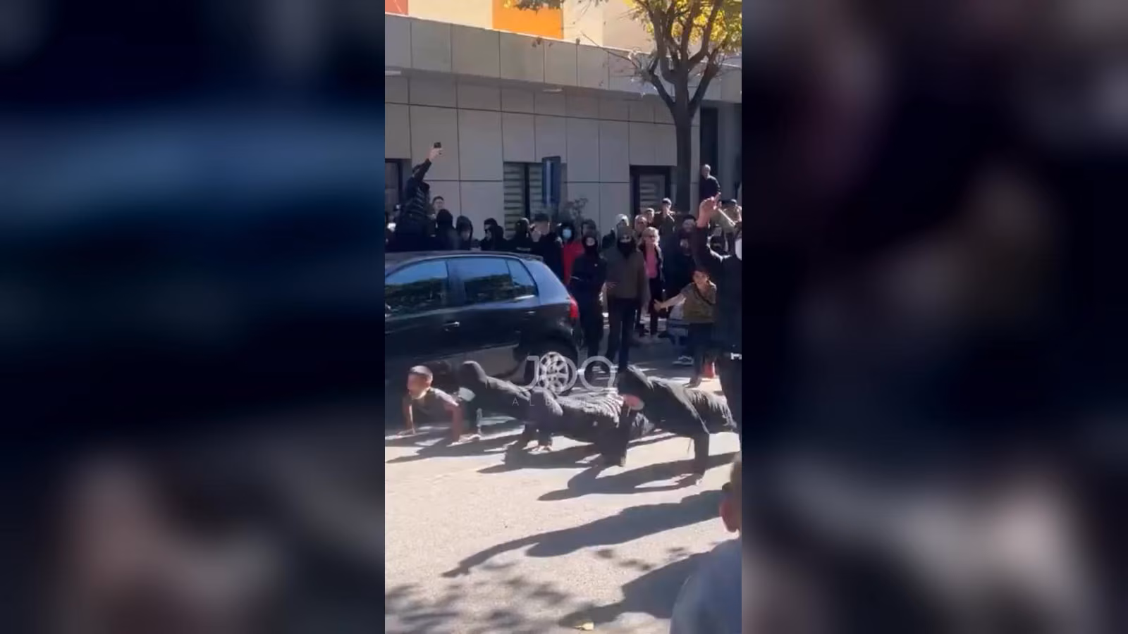 Maskeli, siyah kıyafetli kalabalığın önünde sokakta iki genç şınav çekiyor; yakınlarda siyah bir araba duruyor.