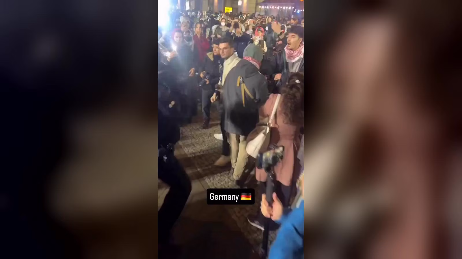 Gece taş döşeli bir caddede toplanan protestocular, birçoğu kırmızı-beyaz kefiye takmış; solda koyu üniformalı biri var.